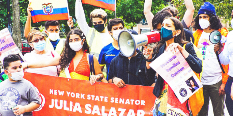 Participants of the Tu Cuentas Immigration March and Census Fair, holding several flags and a red banner with white text that reads "New York State Senator Julia Salazar Senate District 18. The Senator is in the foreground with a megaphone.